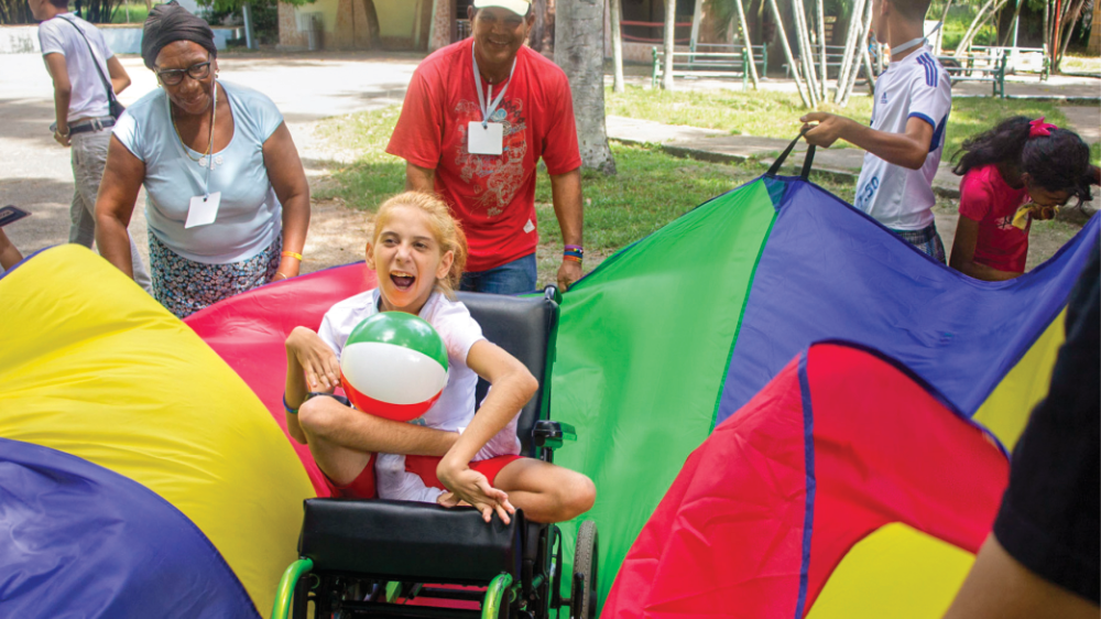 A girl holds a beach ball in her wheelchair while volunteer surround her with a colorful parachute