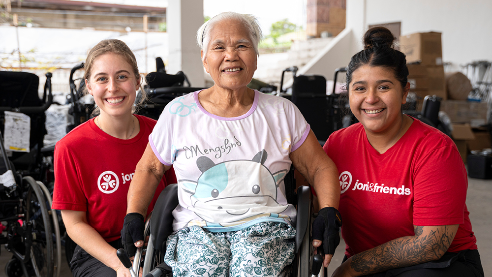 An elderly woman smiles in her wheelchair as volunteer pose for photo next to her