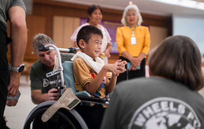 A boy smiles in his new wheelchair as volunteer surround him