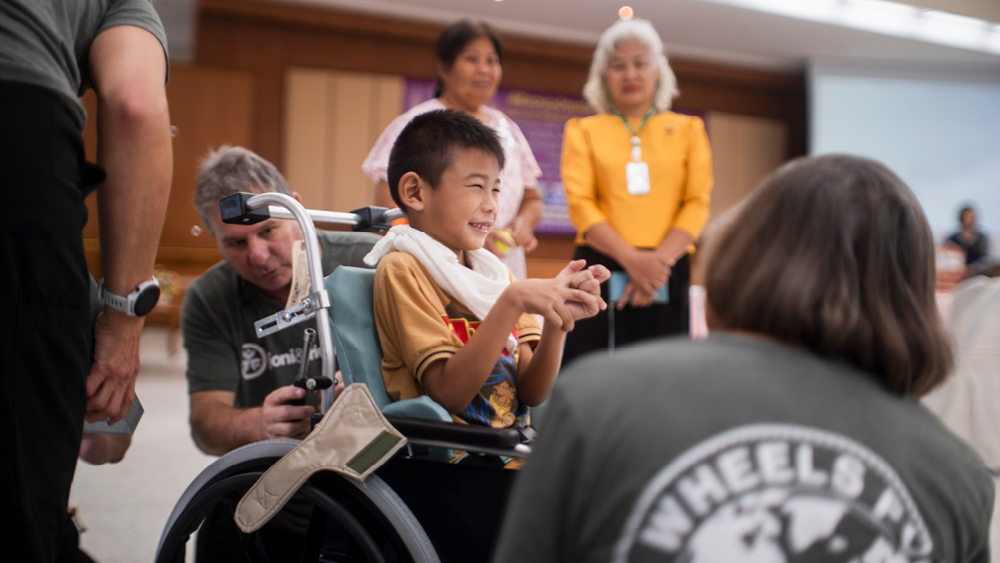 A boy smiles in his new wheelchair as volunteer surround him