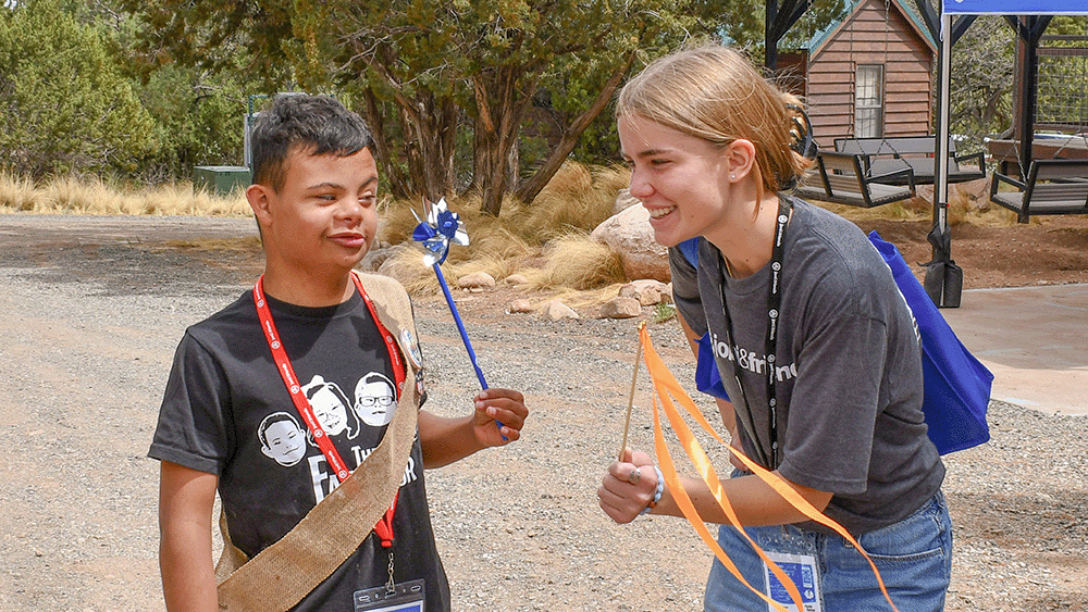 A young boy with a disability holds a pinwheel and smiles at his female volunteer buddy who holds a ribbon on a stick.