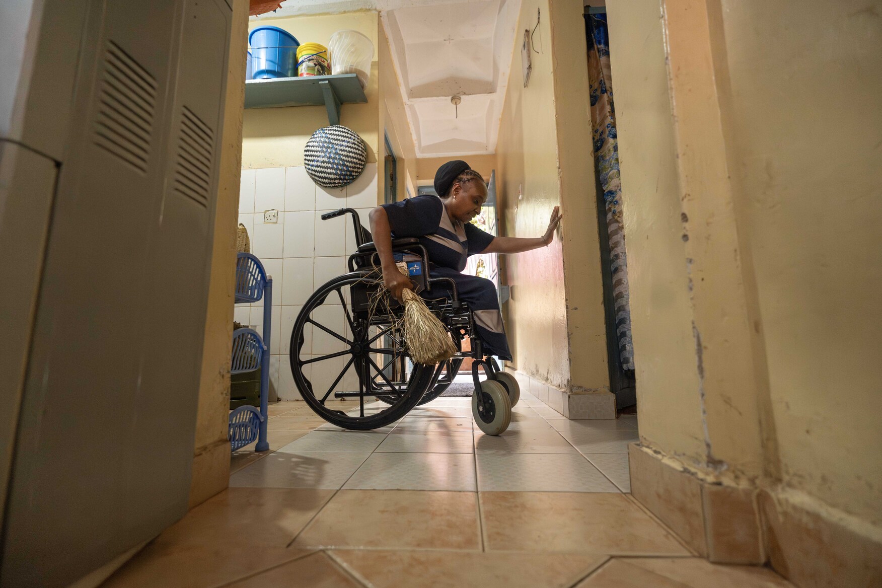 Ruth, sitting in her new wheelchair, holds the wall with one hand and grips a broom with the other.