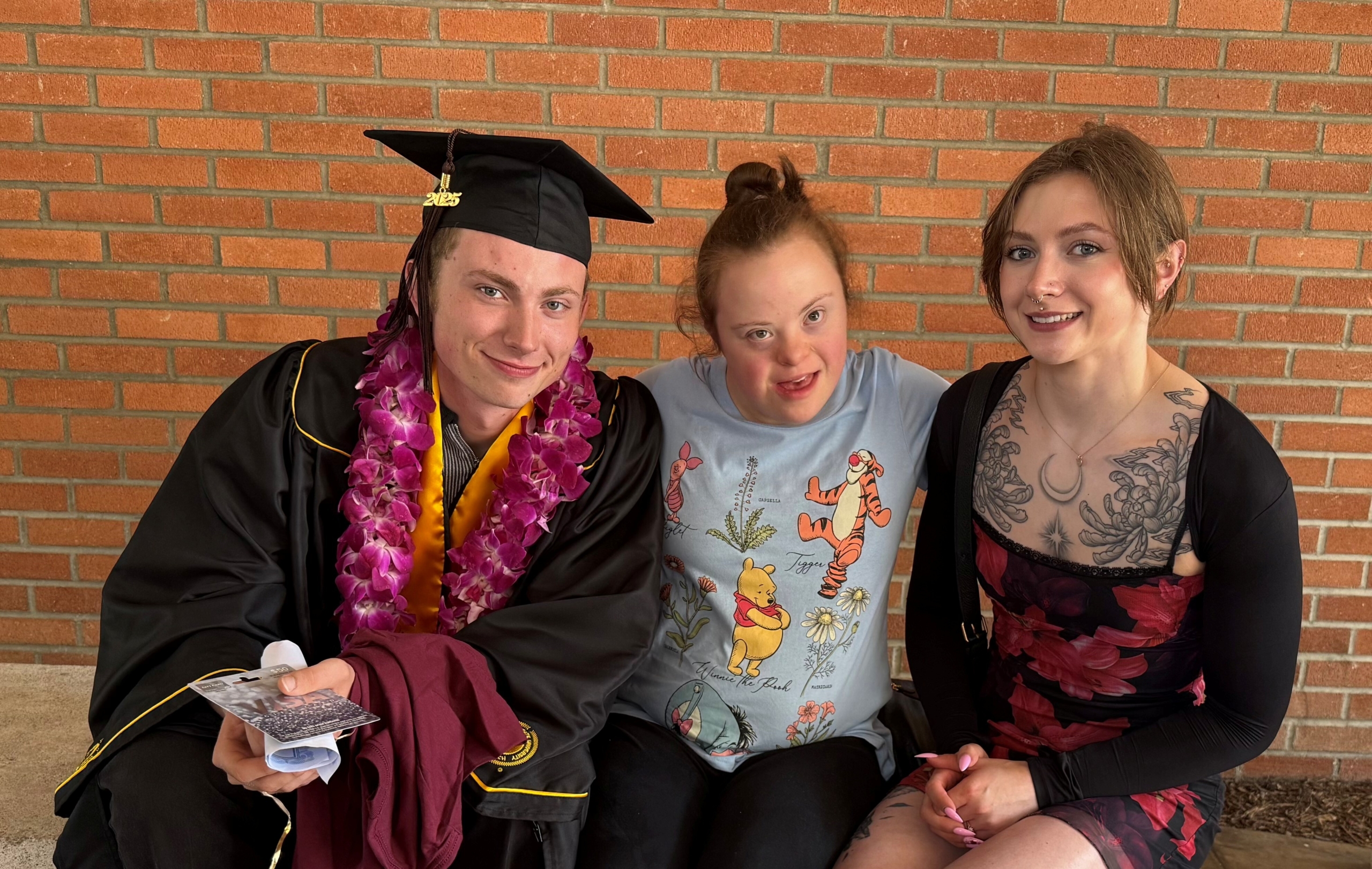 Gideon, wearing his graduation gown and cap, sits on a bench with Sarah and another woman; all three are smiling at the camera.