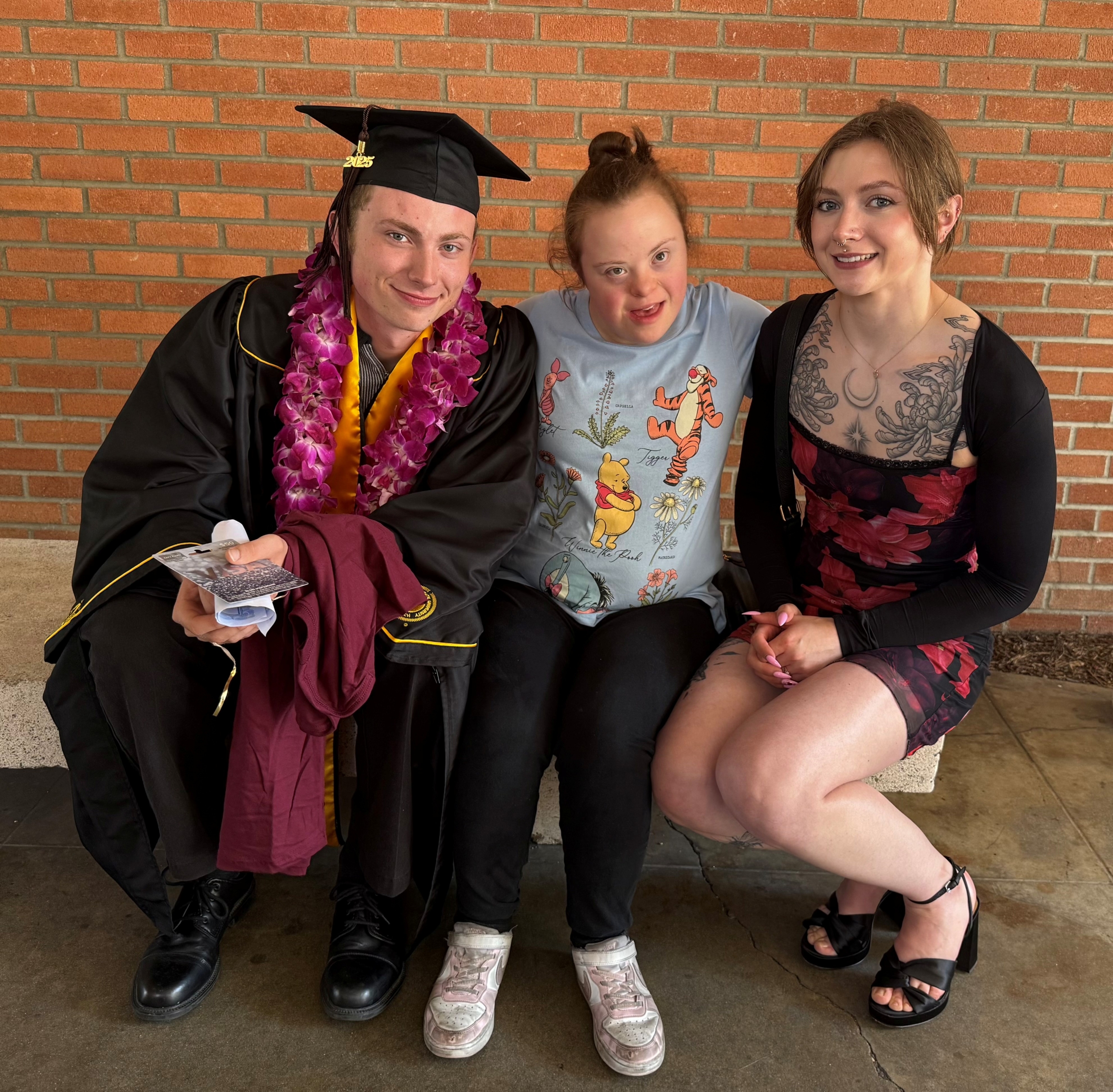 Gideon, wearing his graduation gown and cap, sits on a bench with Sarah and another woman; all three are smiling at the camera.