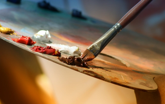 A paintbrush mixing a dark brown paint color on a palette with other colors.