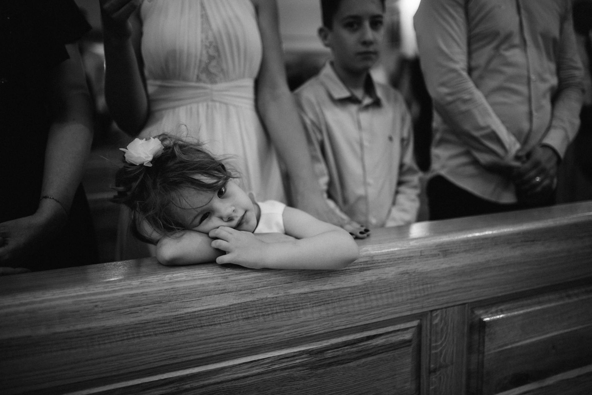 A little girl in a church, looking bored at the camera while resting her head and arm on the wooden pew in front of her.