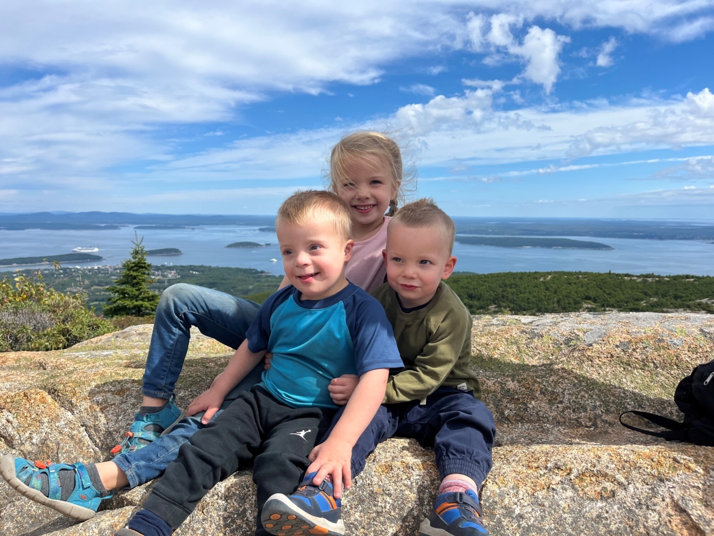 Abigail, Jacob, and Luke sitting on a rock with a view of a body of water and small islands in the background.