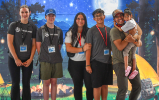 Jayme, Izaiah, and Alexus are carrying Bella, along with two other volunteers, at the Family Retreat. They’re standing in front of a camping-themed backdrop featuring a bonfire, stars, and mountains.