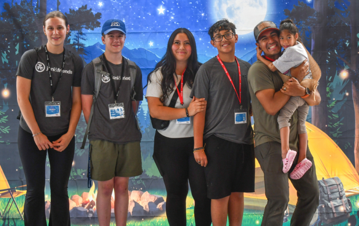 Jayme, Izaiah, and Alexus are carrying Bella, along with two other volunteers, at the Family Retreat. They’re standing in front of a camping-themed backdrop featuring a bonfire, stars, and mountains.