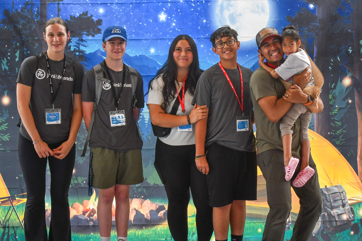 Jayme, Izaiah, and Alexus are carrying Bella, along with two other volunteers, at the Family Retreat. They’re standing in front of a camping-themed backdrop featuring a bonfire, stars, and mountains.