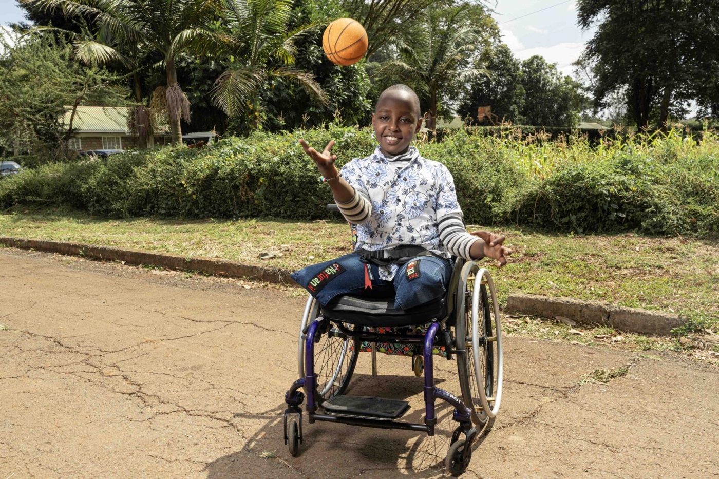 A boy with amputated legs in a sports wheelchair smiles as he tosses a basketball into the air.