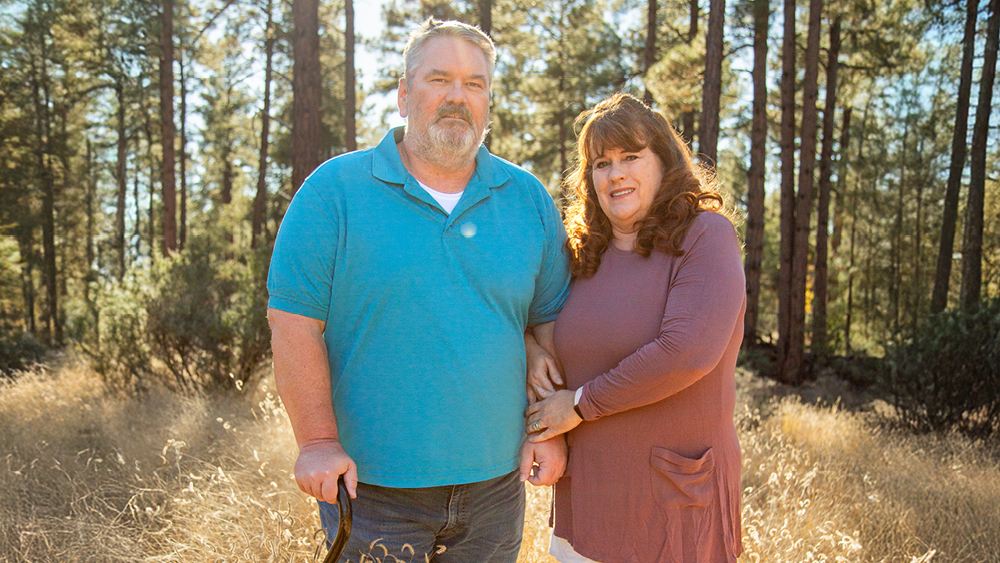 A man standing in front of a forest holds a cane and poses for the camera next to his wife who holds his arm while also posing for the camera.