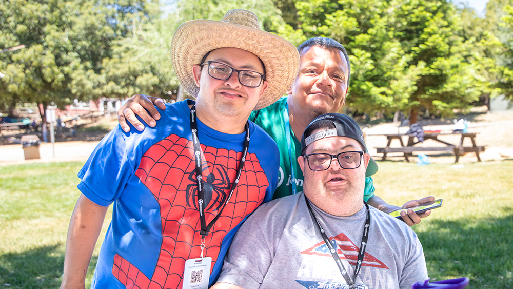 Two men with Down syndrome and their volunteer buddy smile at the Family Retreat volunteer photographer