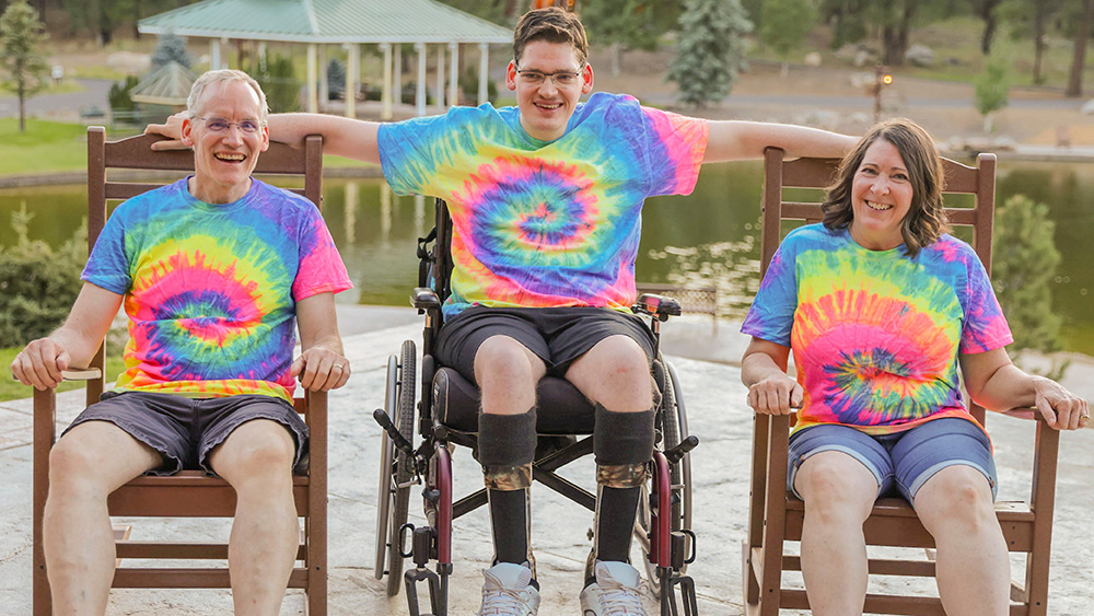 A mother and father sit in rocking chairs on either side of their son who is seated in his wheelchair. They are all smiling at the camera and wearing matching tie-dye shirts.