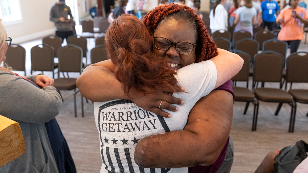 Female Veteran with an amputated left arm hugs another woman wearing a t-shirt that reads "Warrior Getaway" on the back.
