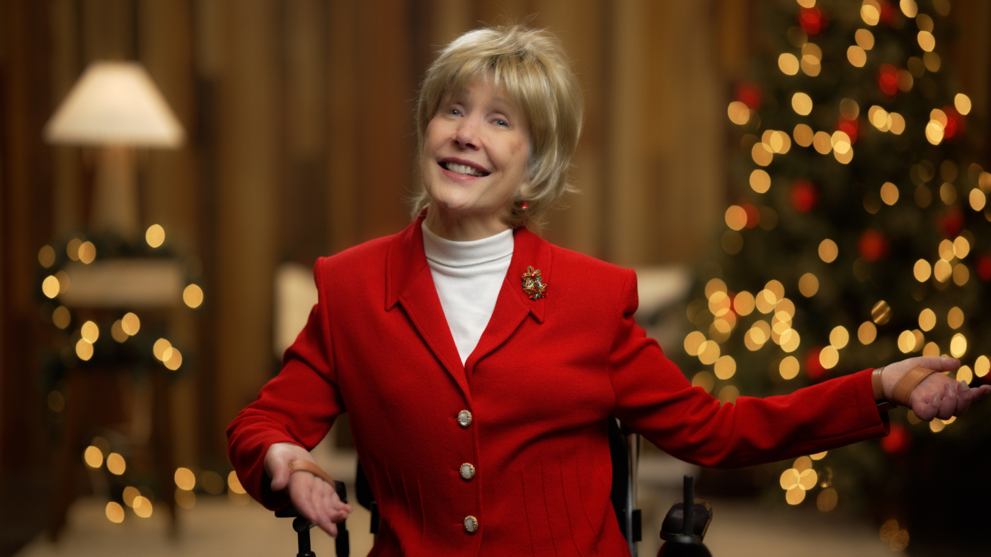 Joni Eareckson Tada smiling for the camera wearing a red blazer with a Christmas bells pin. Behind her are Christmas tree and Christmas lights.