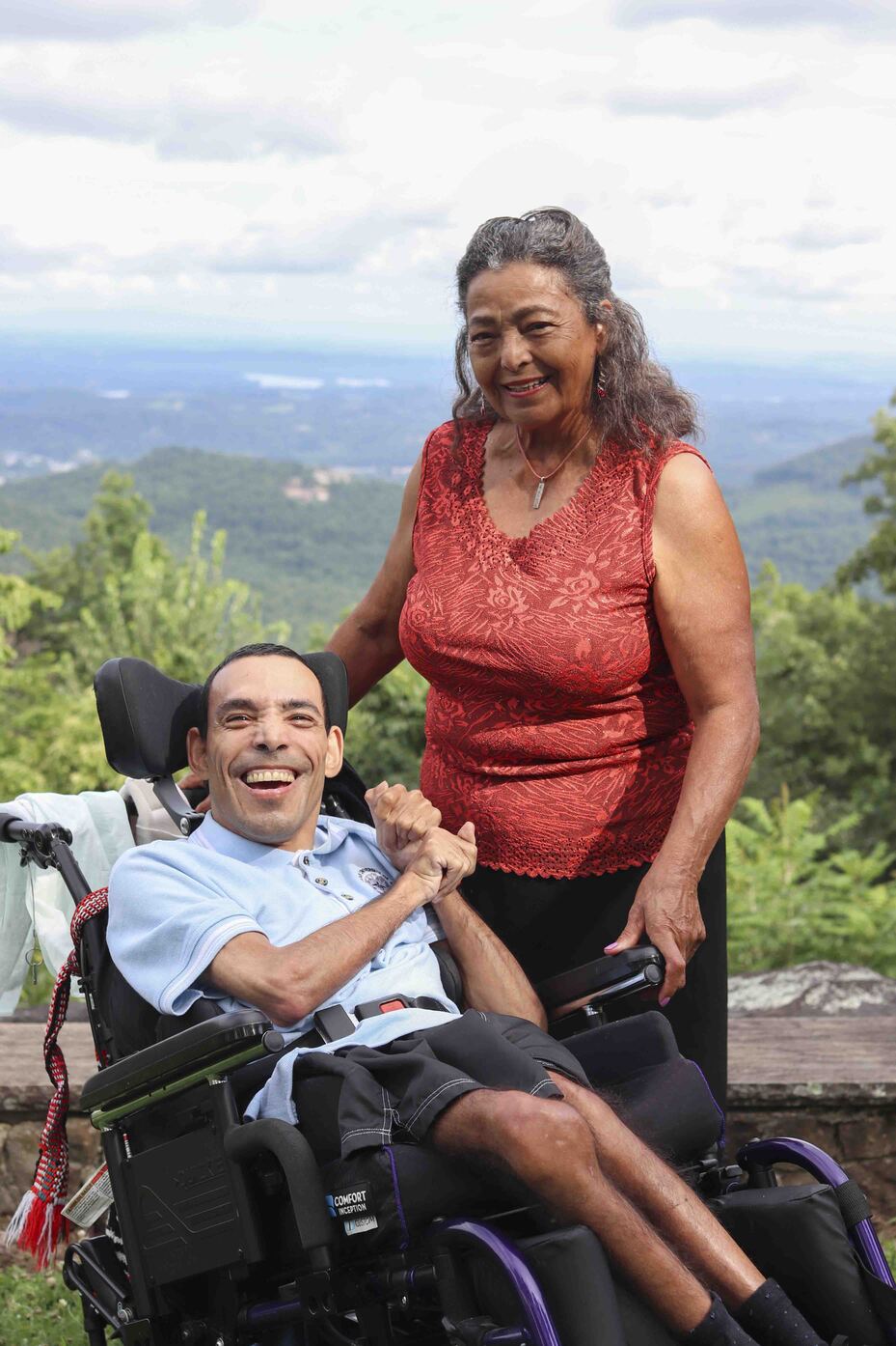 Bruce, seated in his wheelchair, smiles joyfully with a woman standing beside him.