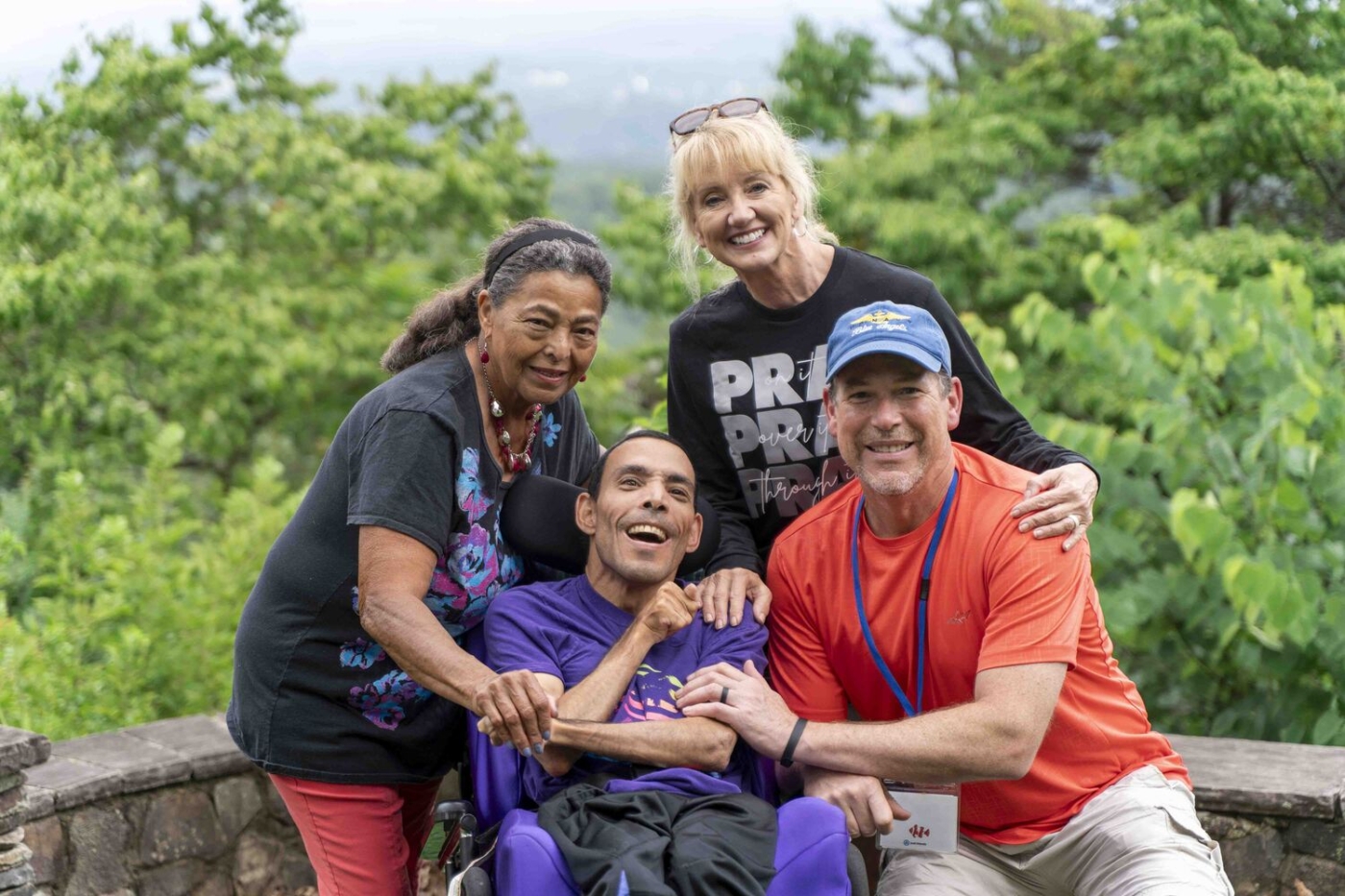 Bruce sits in his wheelchair surrounded by two women and one man, all posing and smiling for the camera.