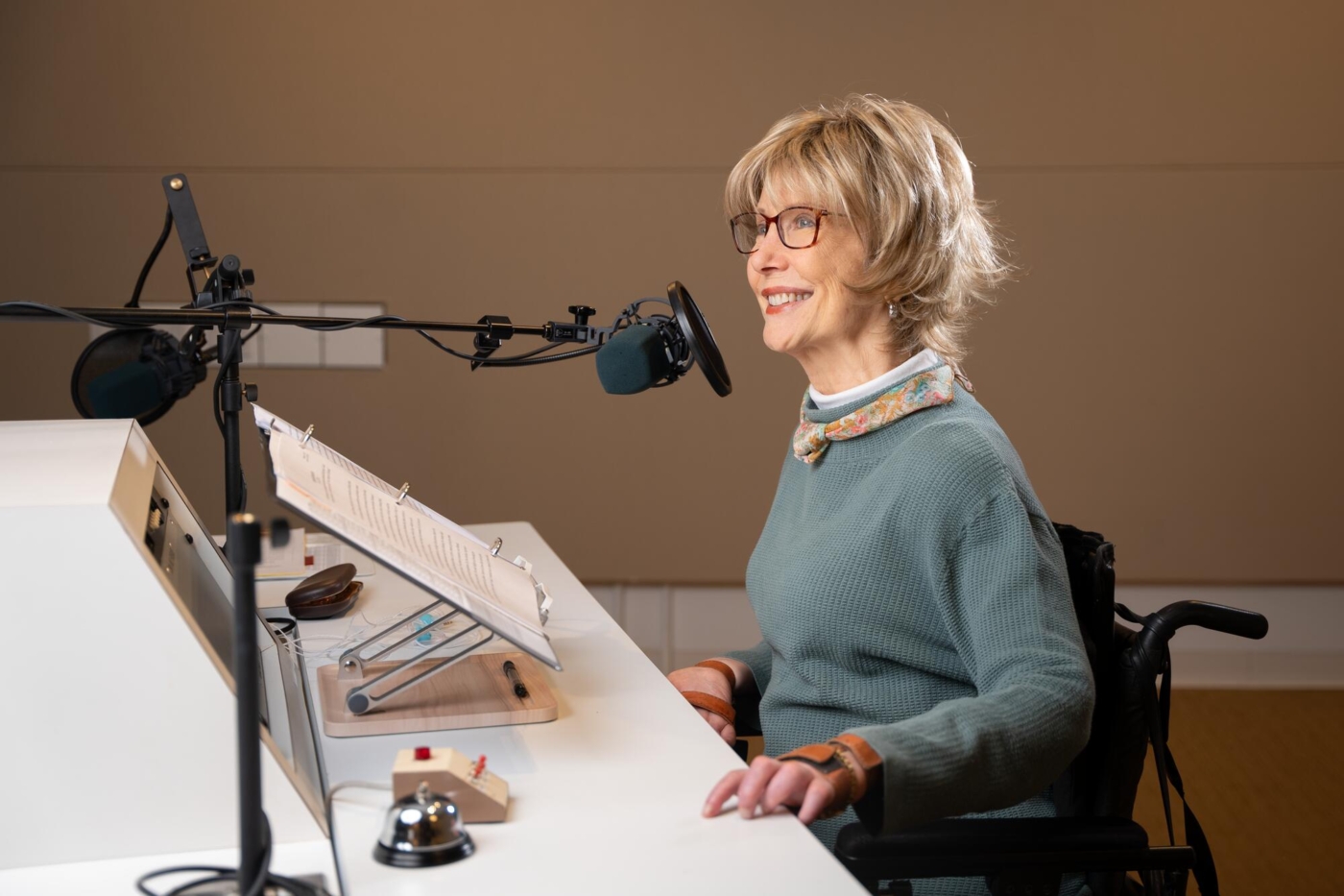 Side view of Joni Eareckson Tada seated in front of a table during one of her radio recordings, speaking into a microphone with a binder containing a script in front of her.