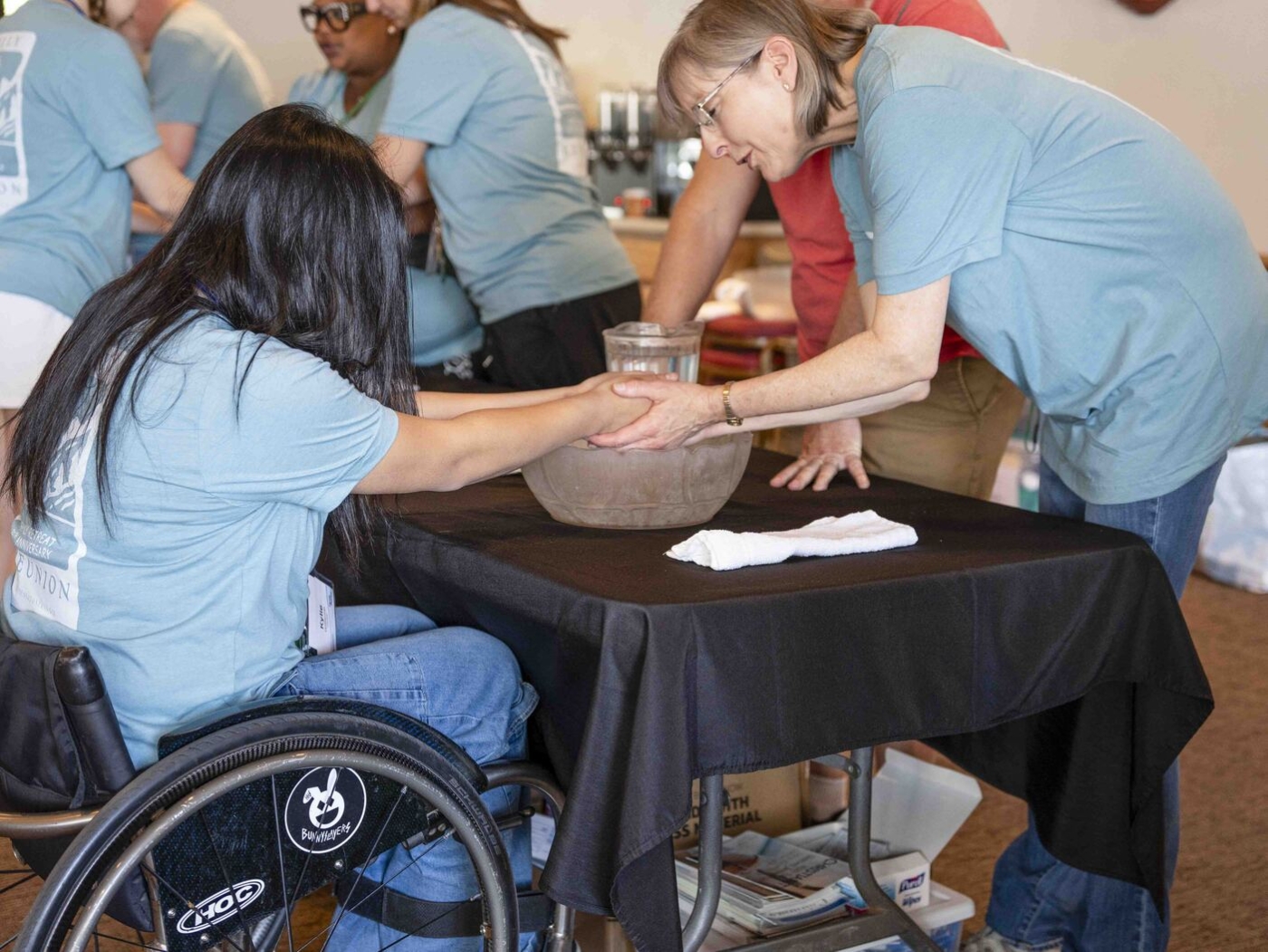 A woman holds the hands of another woman in a wheelchair, with other people visible in the background.