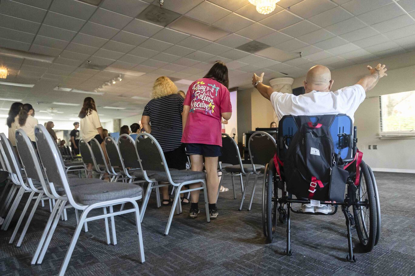 A man in a wheelchair, seen from behind, lifts his arms with other people visible in the background.