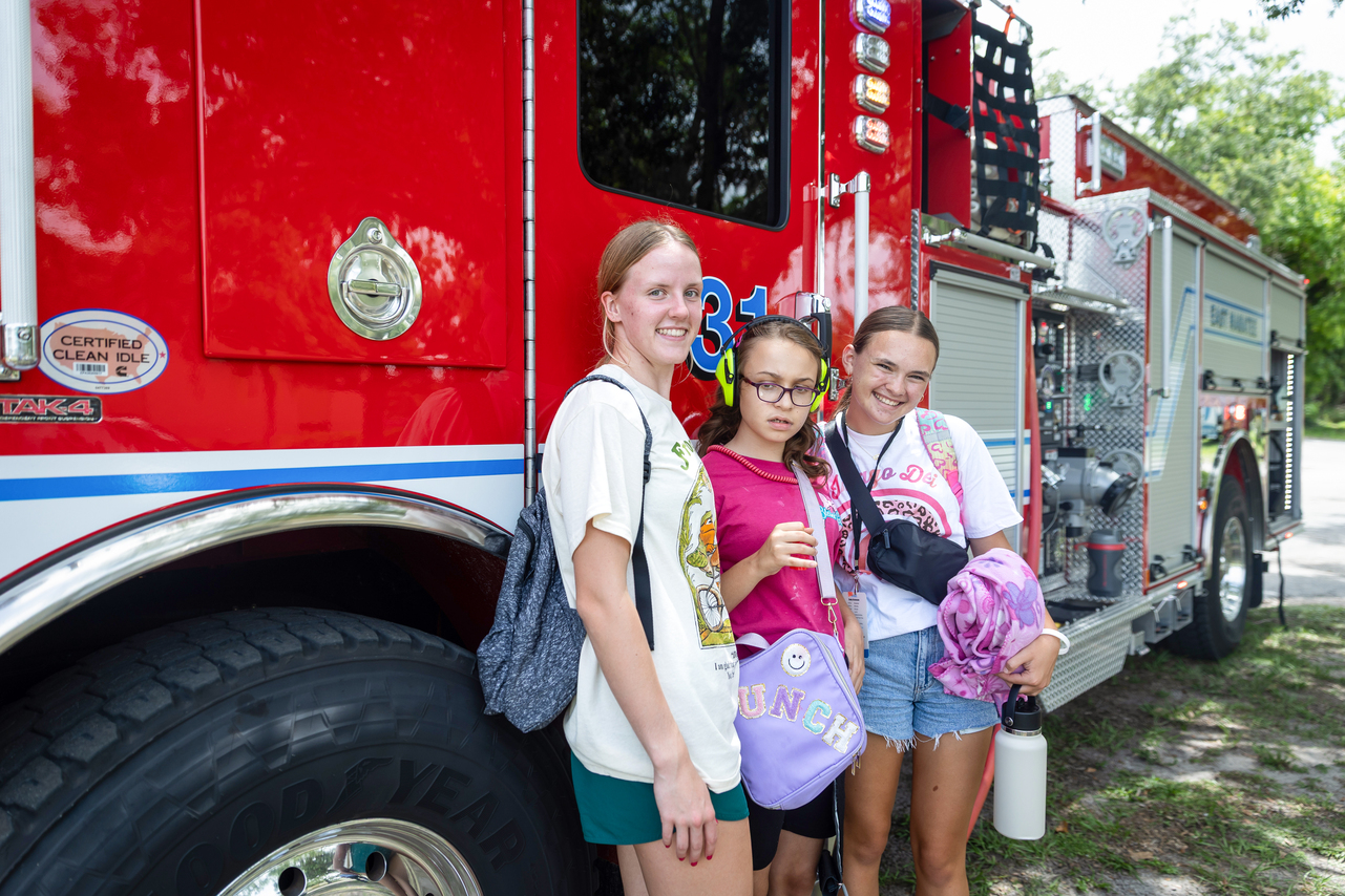 Lindsey wears headphones while standing with two buddies, with a fire truck behind them.