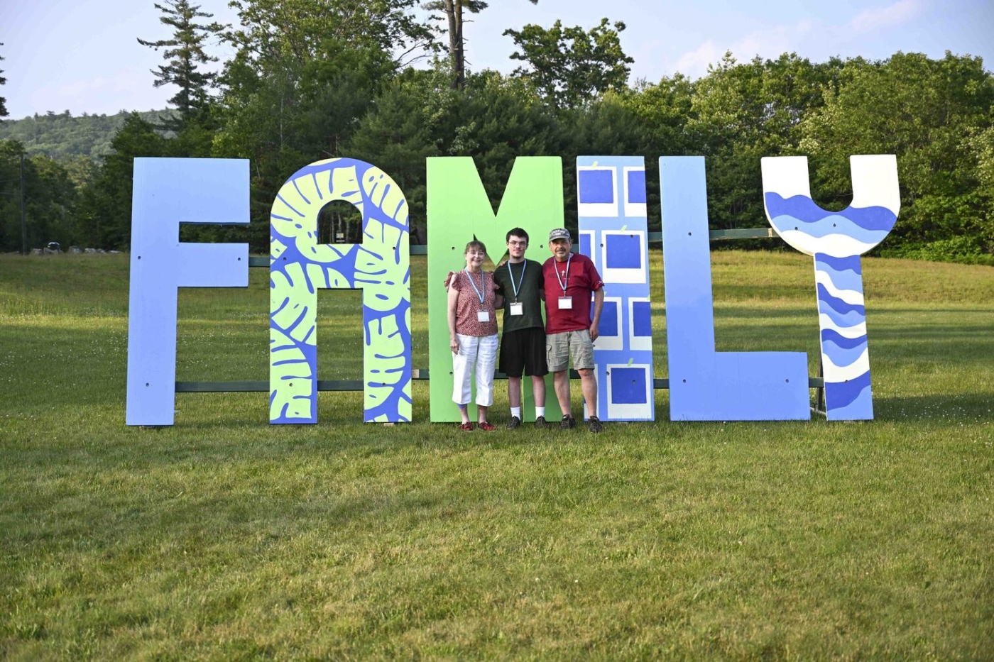 Jim, Annie, and Joshua with a huge stand with the word "FAMILY" behind them standing on the green grass.