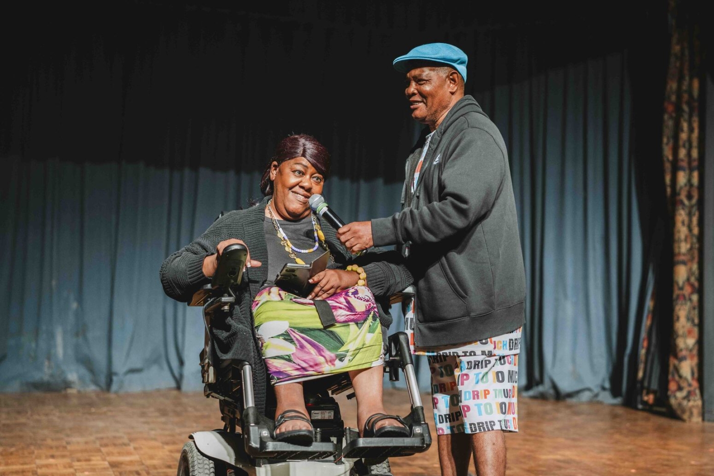 A woman in a power wheelchair speaks into a microphone, assisted by a man standing next to her.