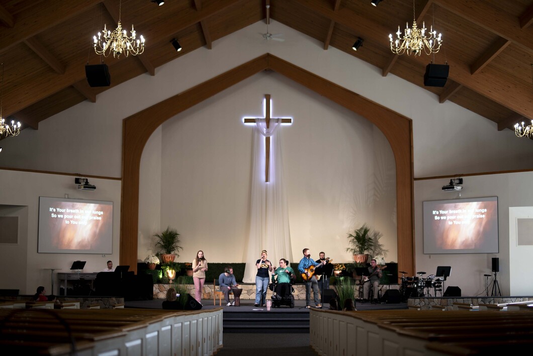 Inside a church, a man in a wheelchair joins others singing during worship, with a large cross displayed prominently in the center.