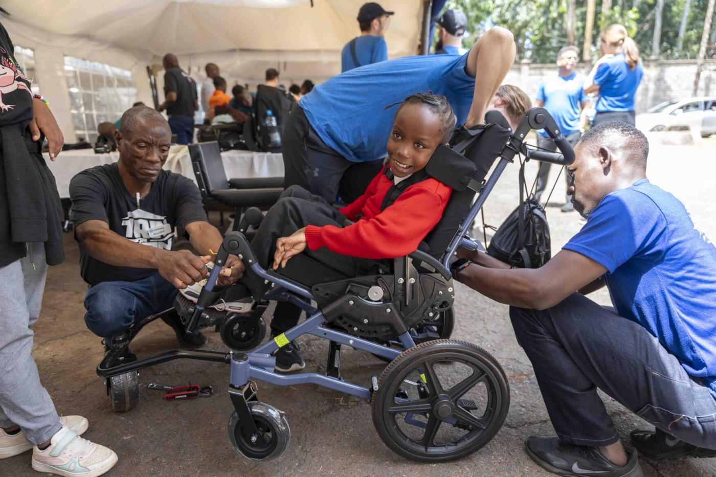 A young boy sits in his new wheelchair, smiling, as men make adjustments to it.