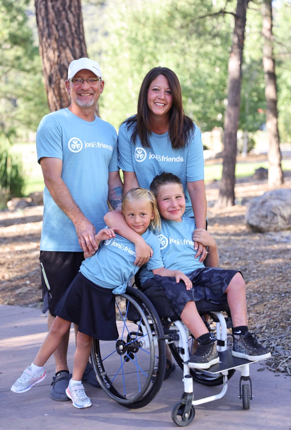 Erin and Charlie stand outdoors with their children, Conrad and Cally, posing for a family portrait. They’re all wearing Joni and Friends T-shirts, smiling with tall trees in the background.