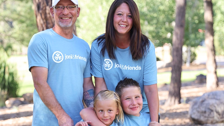 Erin and Charlie stand outdoors with their children, Conrad and Cally, posing for a family portrait. They’re all wearing Joni and Friends T-shirts, smiling with tall trees in the background.
