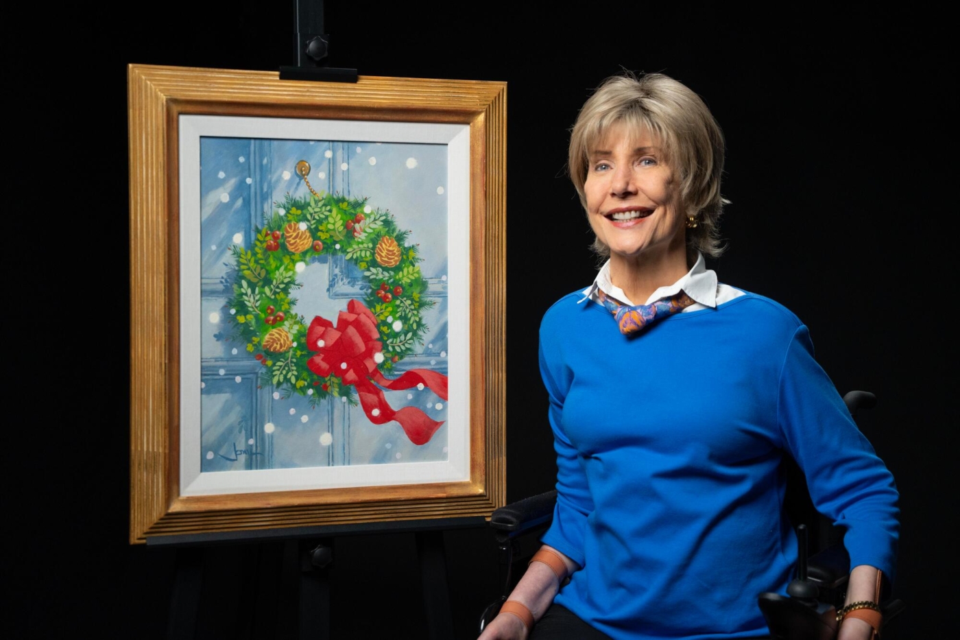 Joni Eareckson Tada smiles beside her artwork titled Christmas Wreath, which features a wreath hanging on a door with snowfall.