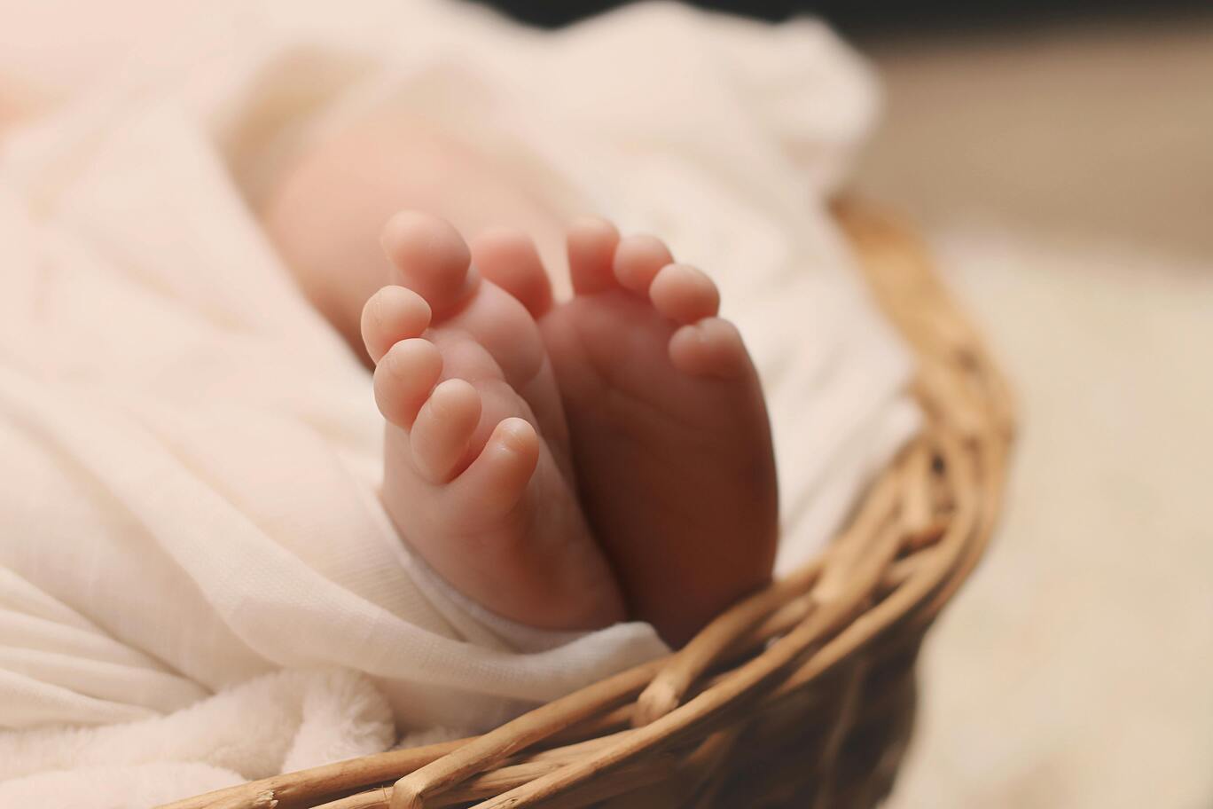 A baby wrapped in white linens, showing tiny feet, lying in a woven basket.