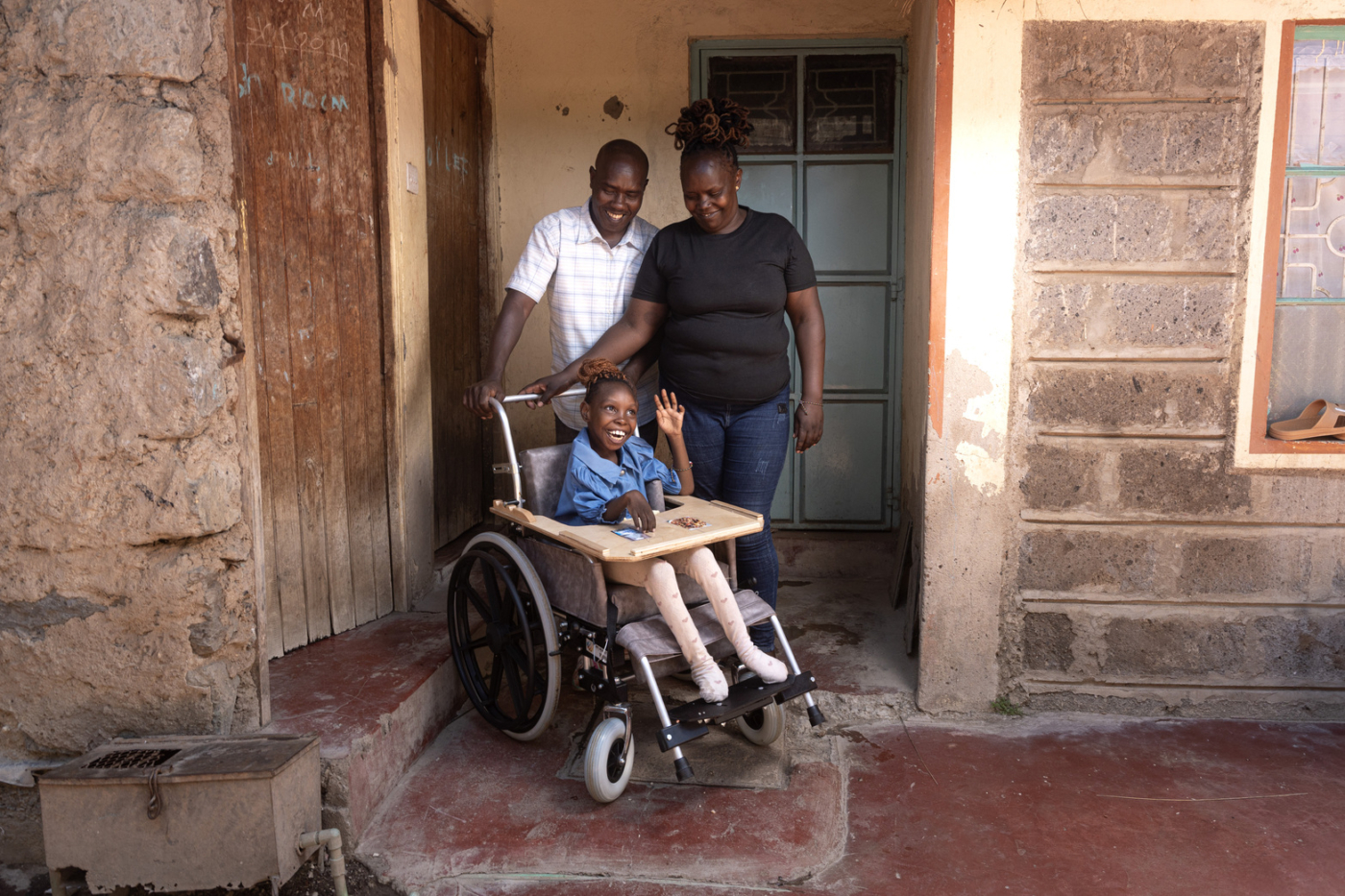 Praise sits in her new wheelchair and waves while her mother and father stand behind her, looking at her.