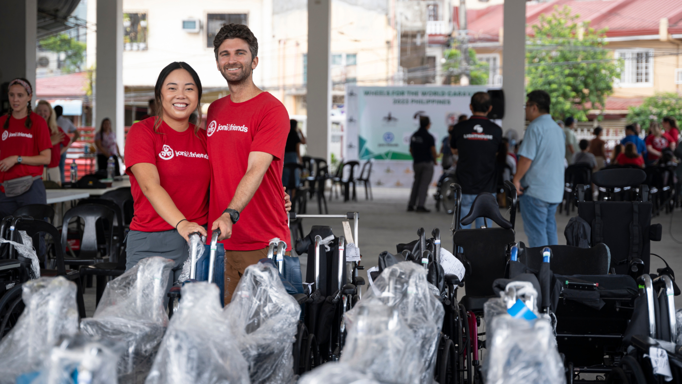 A husband and wife stand together, smiling at the camera, and holding the handles of wheelchairs wrapped in plastic wrap waiting to distribute them to people with disabilities.
