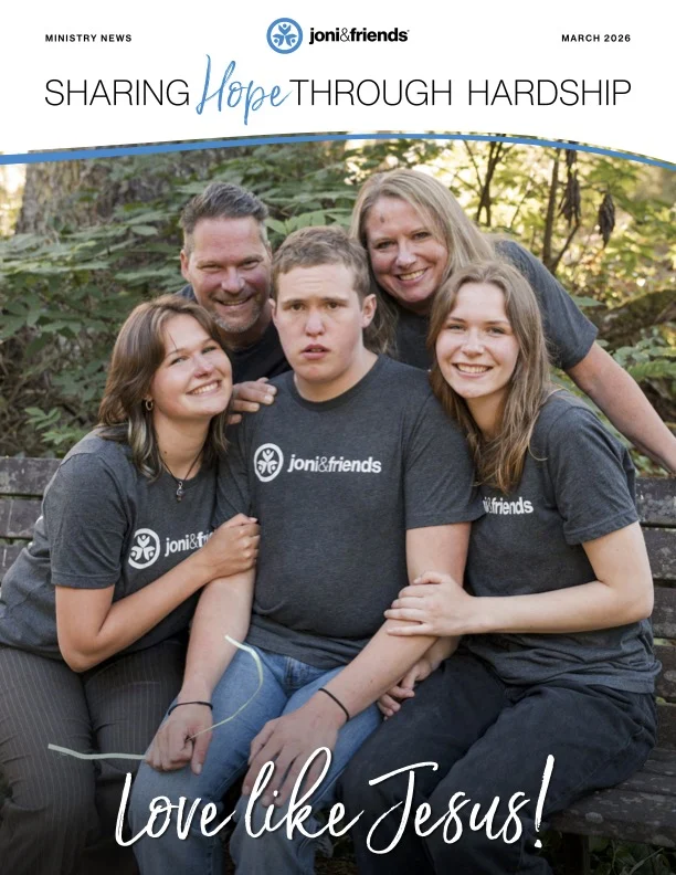 A man with a disability sits in front, smiling at the camera, with his two sisters beside him and his parents standing behind him. Everyone is smiling and posing for the photo.