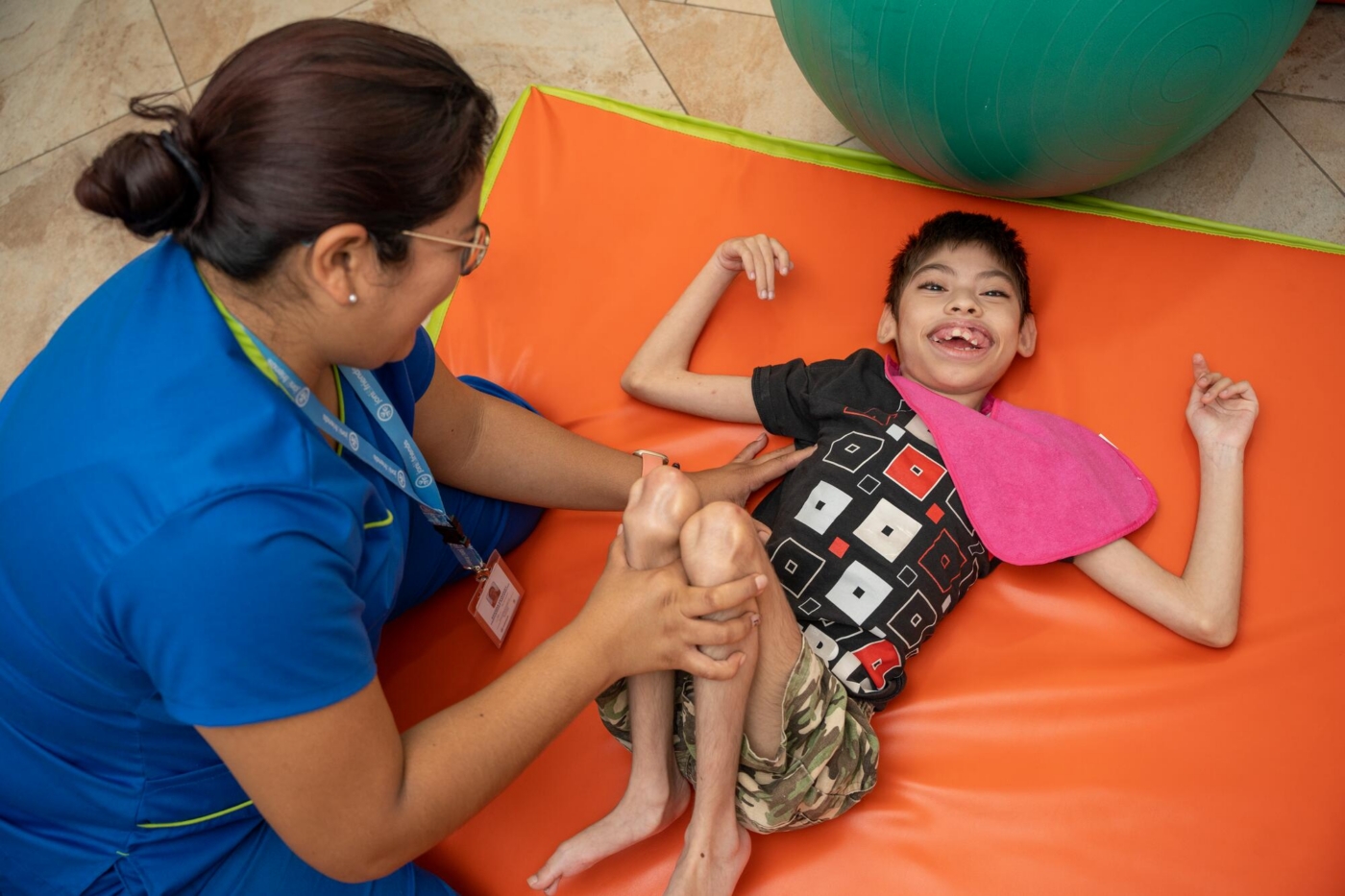 A boy lying on a mat smiling at the camera while a physical therapist stretches him.