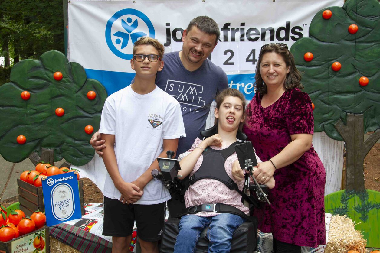 Sophia sits in her wheelchair with her mom, dad, and another boy in front of a Joni and Friends backdrop decorated with trees and oranges.