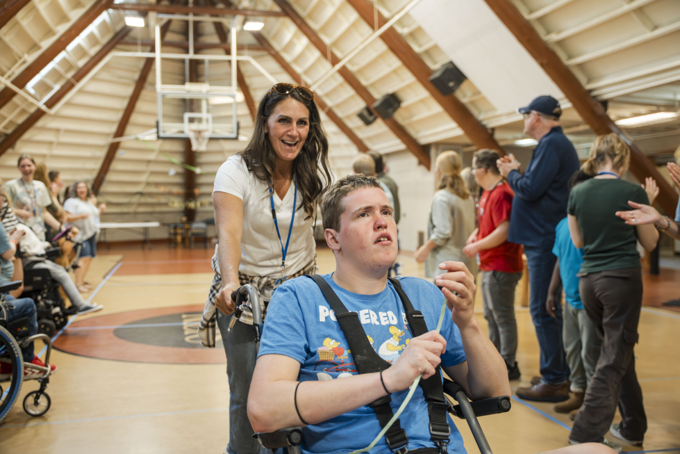 Simon sitting in his wheelchair with a volunteer behind him, pushing him while smiling.