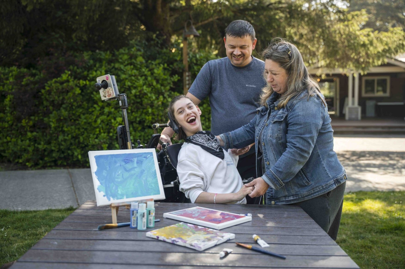 Sophia sits in her wheelchair smiling at the camera while her parents stand in front of her. A table with canvas and paint is set up nearby.