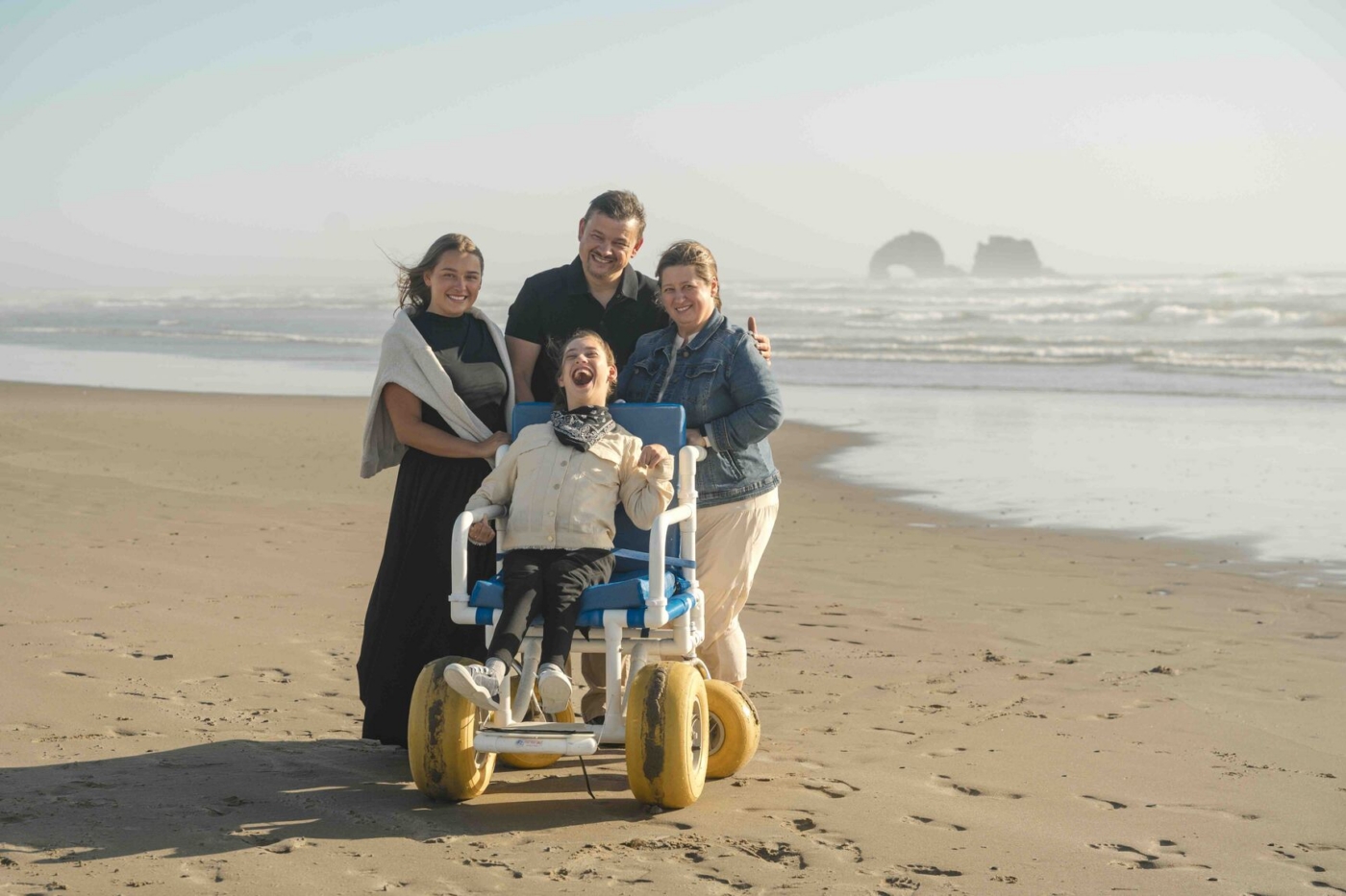 Sophia sits in a beach wheelchair with her father, mother, and another woman by the shore.