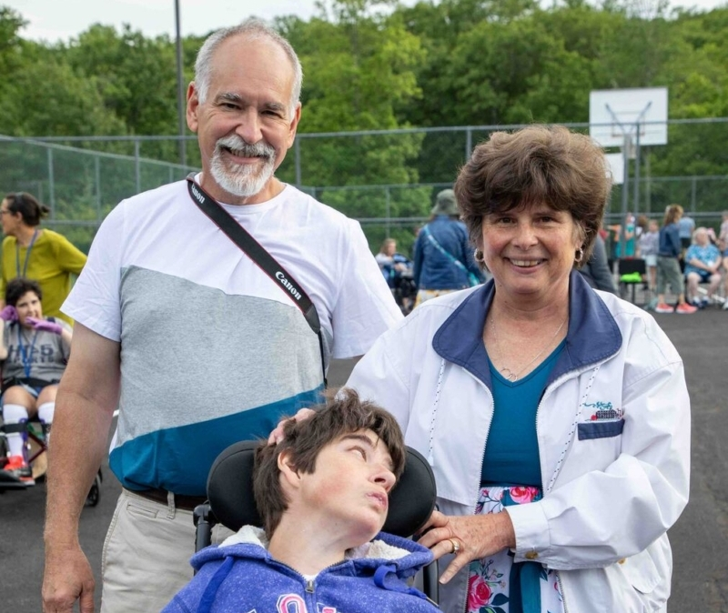 Kari sitting in her wheelchair with her parents Ron and Joan standing behind her, smiling at the camera.