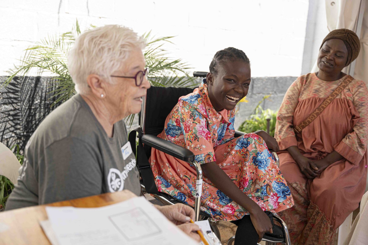 Mapalo smiles at the camera while sitting in a wheelchair beside volunteer Rose and her mother, Kongwa.