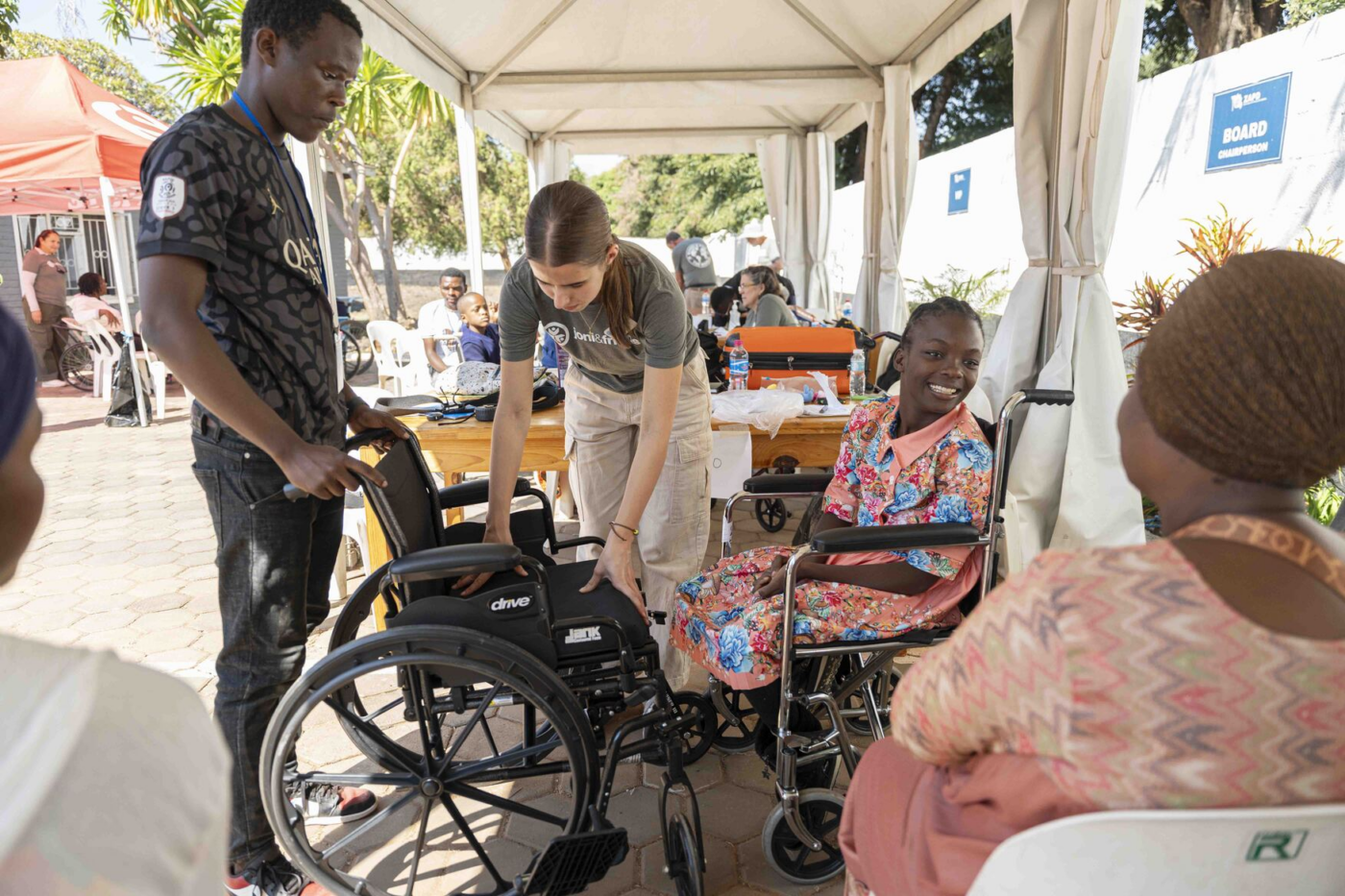 Mapalo sits in her wheelchair, looking at her mom while volunteers in front of them are holding another wheelchair.