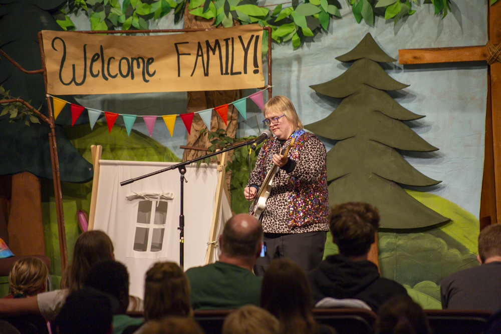 Emily, a woman with Down syndrome, plays an electric guitar and sings into a microphone on stage while an audience watches.