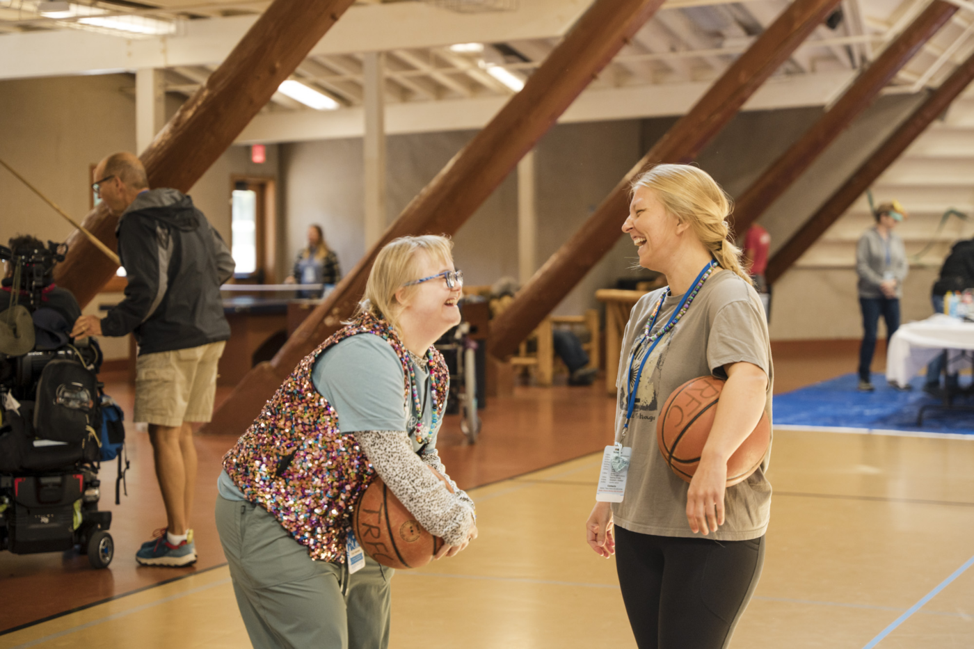Emily, a woman with Down syndrome, laughs with her buddy Katie while they hold a basketball on an indoor court.
