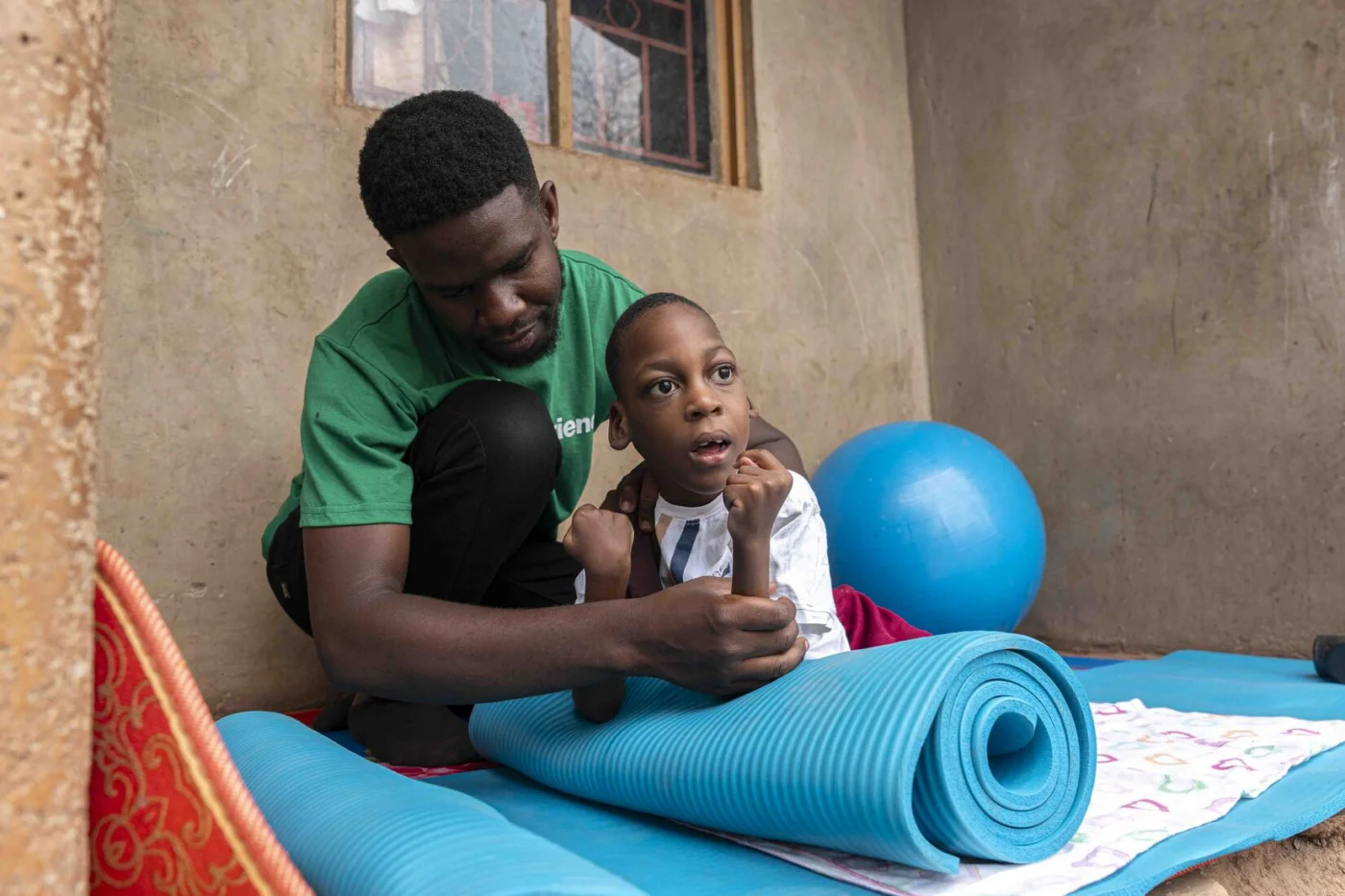 A man assisting Favour during a physical therapy session on a mat, with a stability ball positioned behind them.