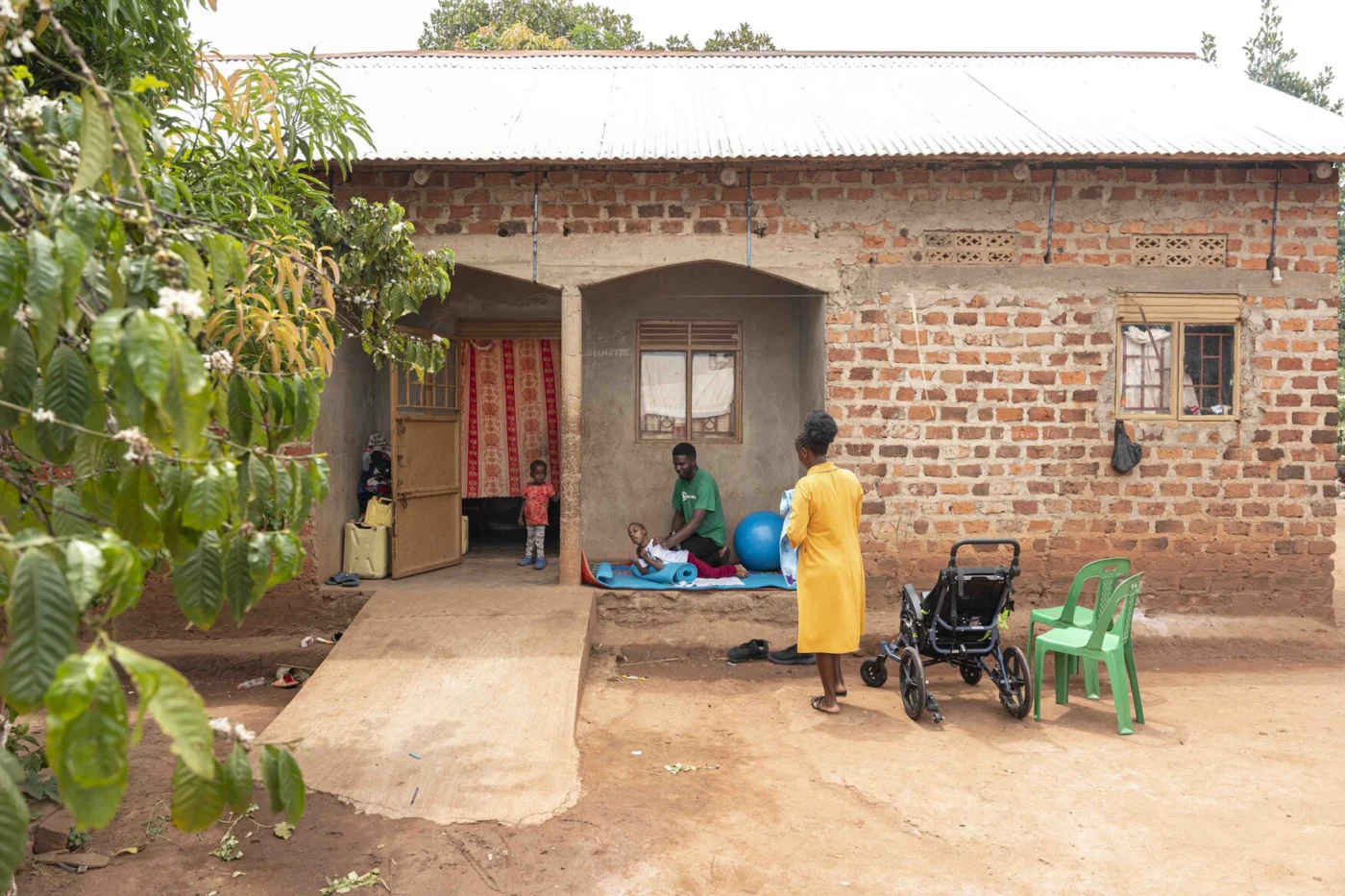 Overview of Favour's house showing a ramp. Favour is doing physical therapy on a mat with a man assisting him. His mom stands nearby next to his new wheelchair, and his little brother watches from the doorway.