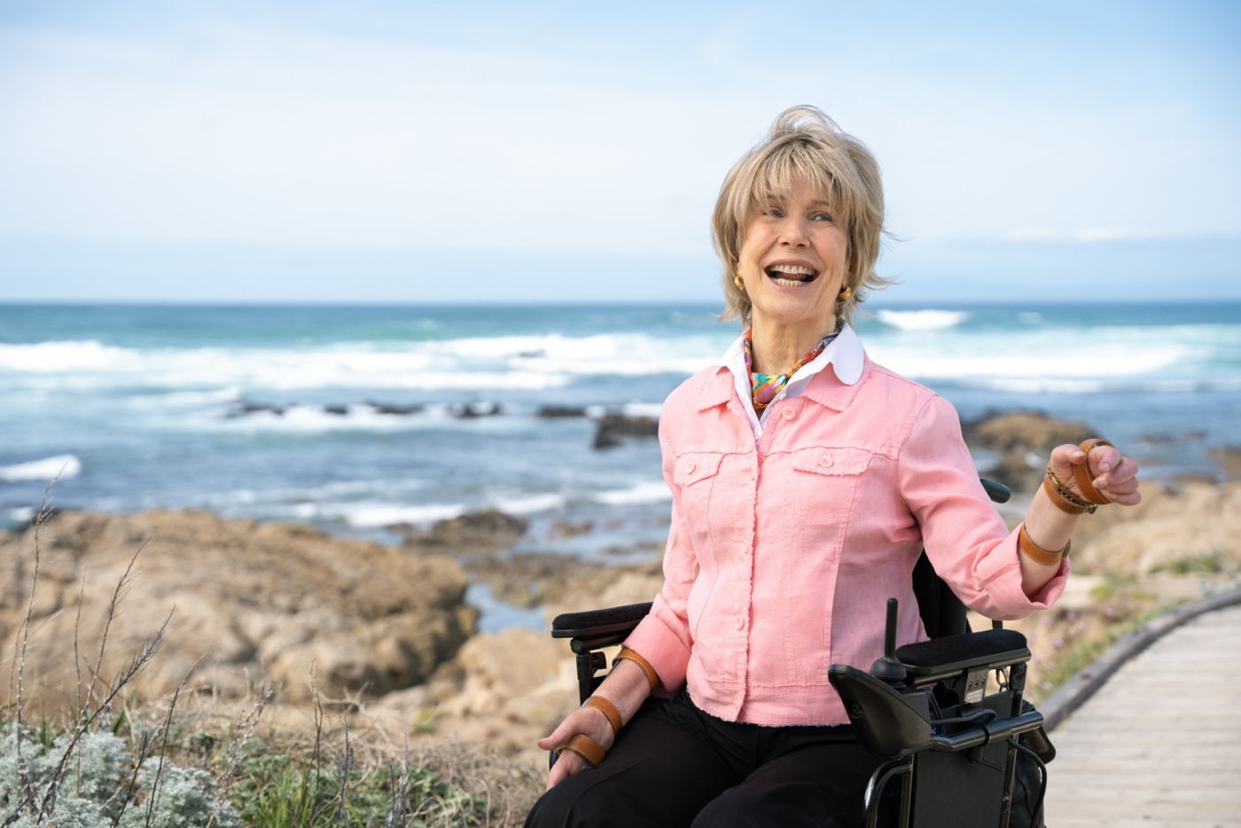 Joni sitting in her wheelchair with wind blowing her hair, with ocean waves in the background.